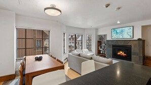 A modern living room with a fireplace, a dining area, and large windows offering a view of buildings.