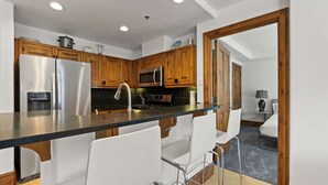 A modern kitchen with wooden cabinets, stainless steel appliances, and a bar area with white chairs.