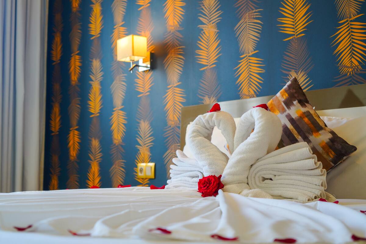 A neatly made bed with towels and a rose, a wall with a leaf pattern, and a wall-mounted lamp.