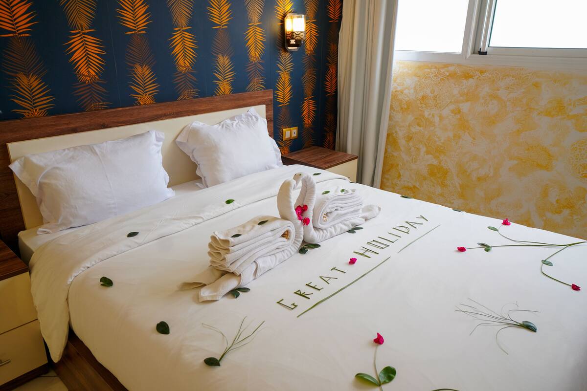 A neatly made bed with towels and a floral bedspread, a wooden headboard, and a wall with a leaf pattern.