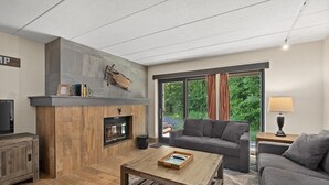 A modern living room with a fireplace, grey sofas, a wooden coffee table, and a large window with a view of greenery.