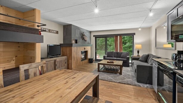 A modern living room with a wooden dining table, a sofa, and a kitchen area with a TV and coffee machine.