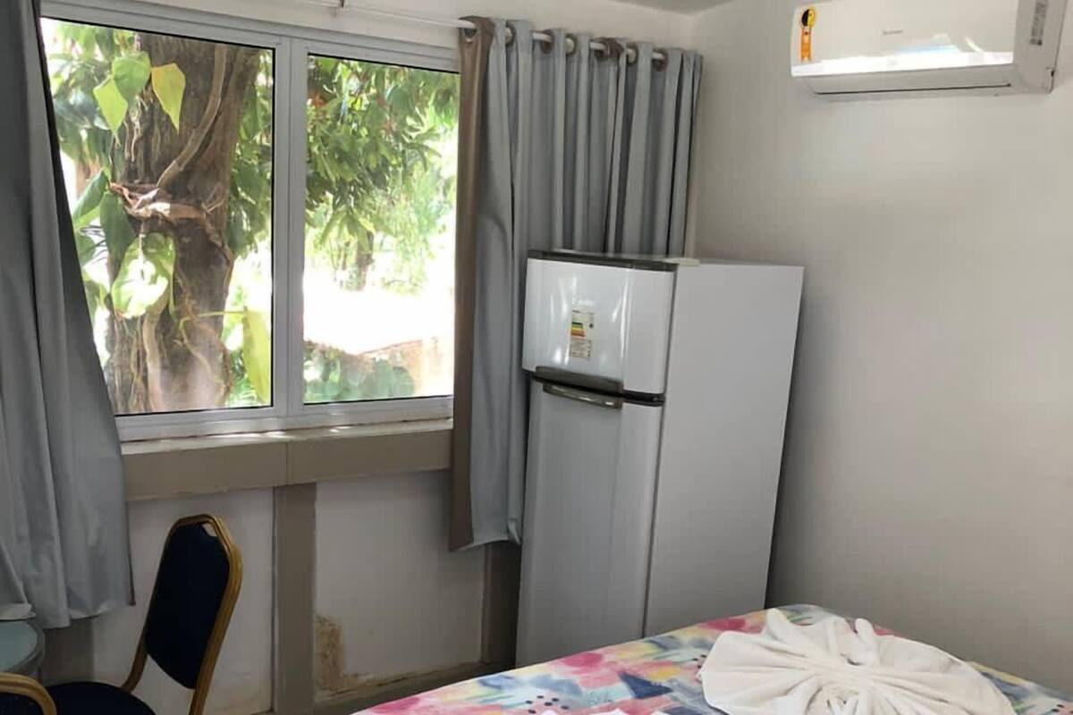 A hotel room with a bed, a chair, a window with curtains, a refrigerator, and a wall-mounted air conditioner.