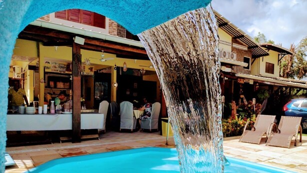 A swimming pool with a waterfall feature, outdoor seating, and a building with a covered patio.