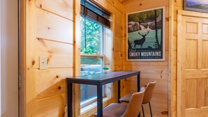 Et interiør af en træhytte med et bord og to stole, et plakatbillede af Smoky Mountains, og et vindue med udsigt til grøn vegetation.