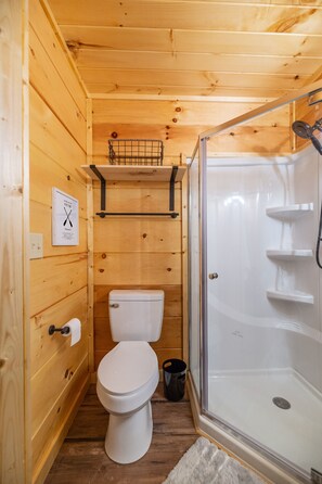 A wooden cabin bathroom with a toilet, a shower enclosure, and a shelf above the toilet. - Tiny Cabin -Built in 2022- with Amazing View and Hot Tub! (Sevierville)
