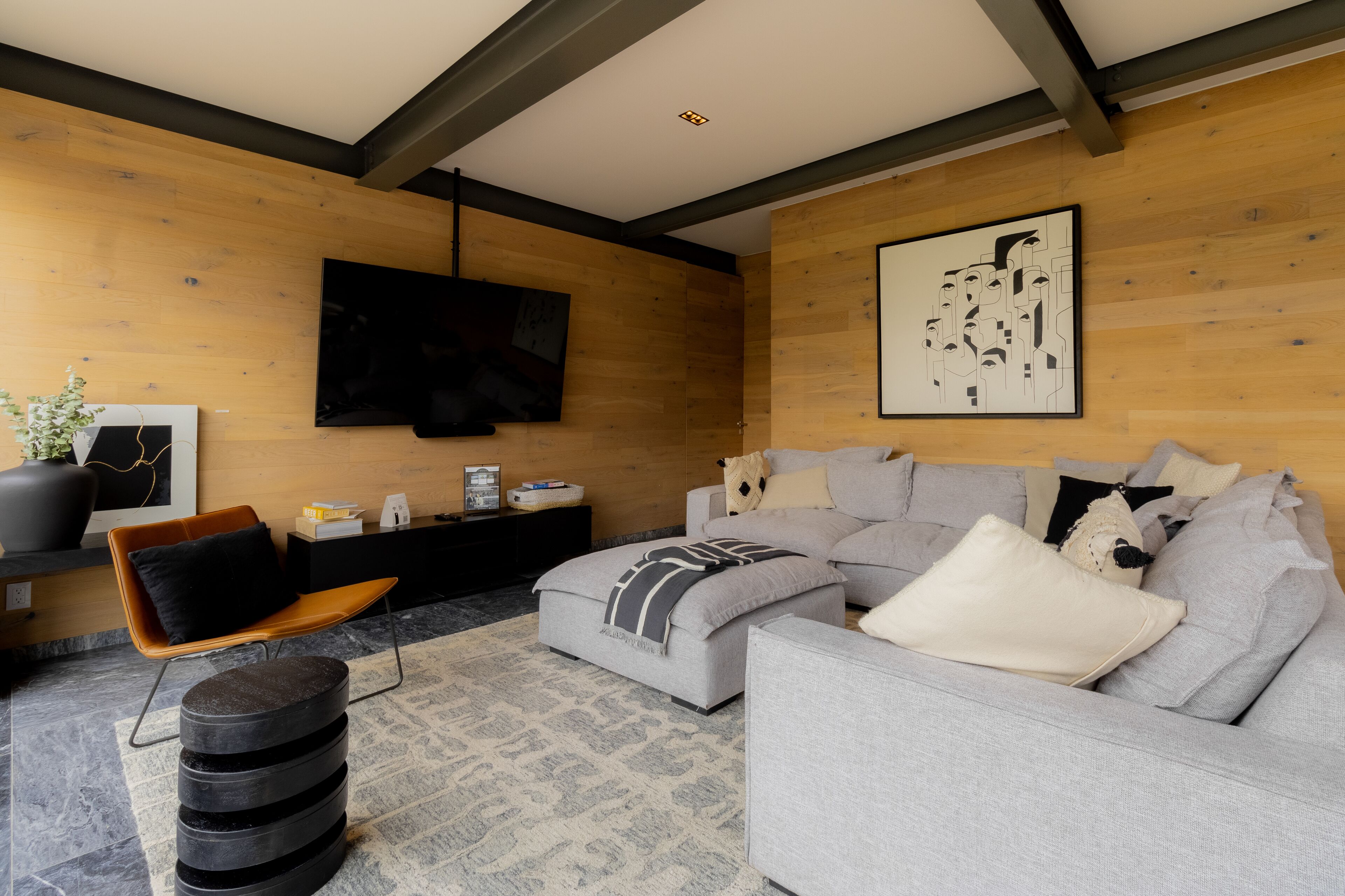 A modern living room with a grey sectional sofa, a black ottoman, a wooden accent wall, a mounted flat-screen TV, and a framed abstract art piece.