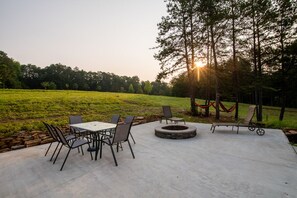 An outdoor patio area with a round table and chairs, a fire pit, and a view of a grassy field and trees. - The Jocassee Tiny House (Salem)