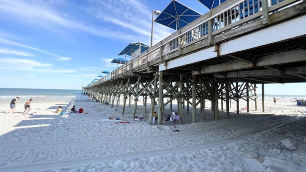 A beach with a wooden pier extending into the ocean, featuring blue umbrellas and a railing. - Sand In My Boots<br>Condo Beach view, 3 bed, 1 bath (Murrells Inlet)
