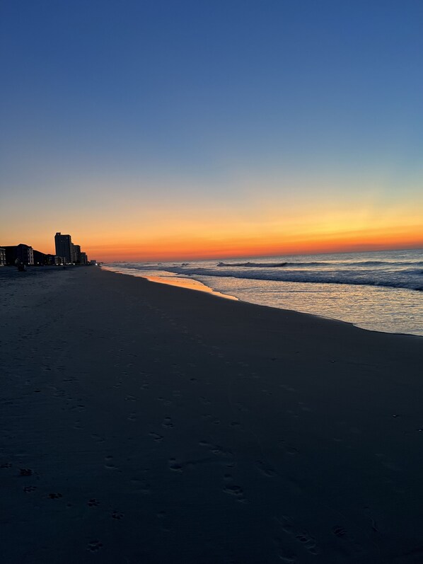 A beach with footprints leading to the water, a clear sky, and buildings in the background. - Sand In My Boots<br>Condo Beach view, 3 bed, 1 bath (Murrells Inlet)