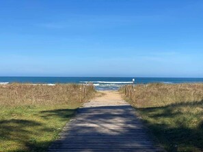 En træpromenade fører til stranden, omgivet af græsplæner.