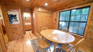 A wooden cabin interior with a dining table, chairs, a rug, and a large window offering a forest view. - Wolf`s Den - Dogs OK- Hot Tub-Firepit-Wi-Fi-Fireplace-Covered Deck with Gas Grill (Blue Ridge)
