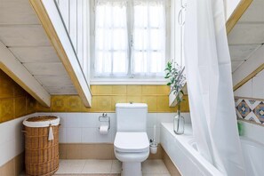 Un baño pequeño con inodoro, una cesta de mimbre, un dispensador de papel higiénico, un jarrón de vidrio con una planta y una bañera con cortina blanca.