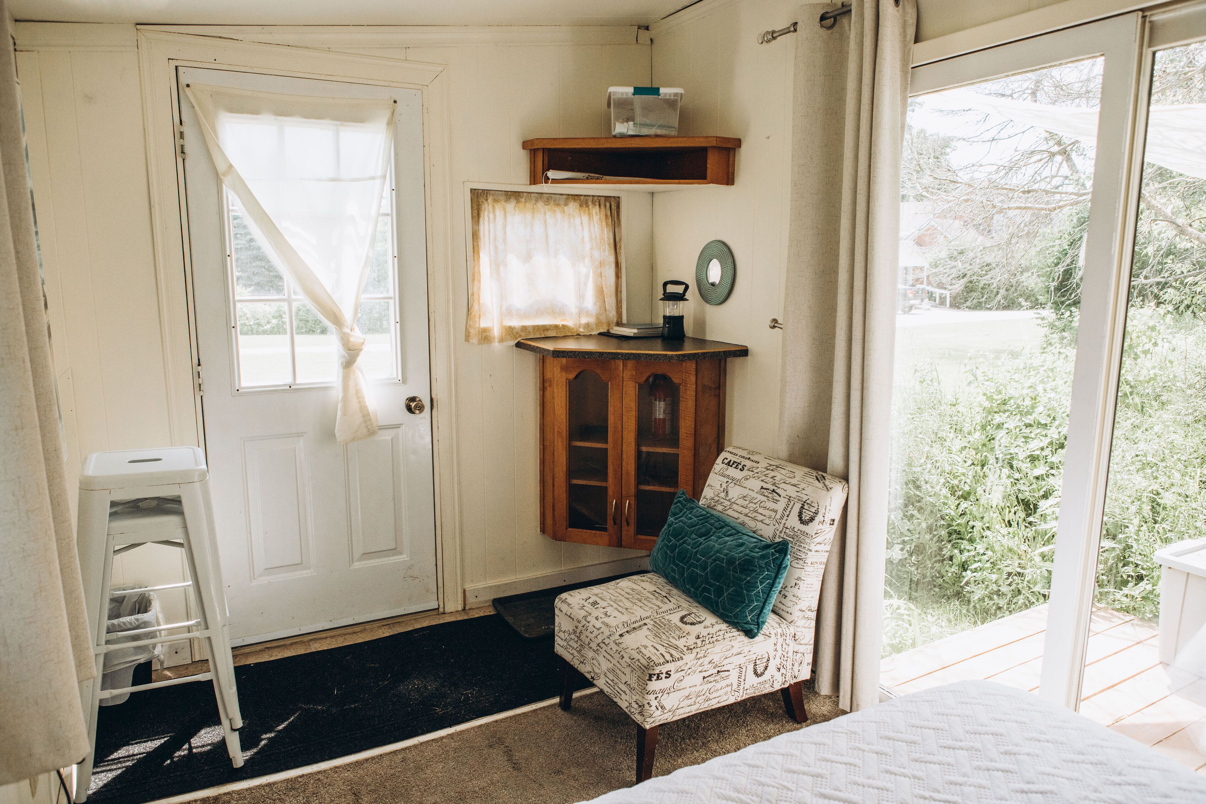 A room with a bed, a chair with a cushion, a wooden cabinet, a white door, and a window with curtains.