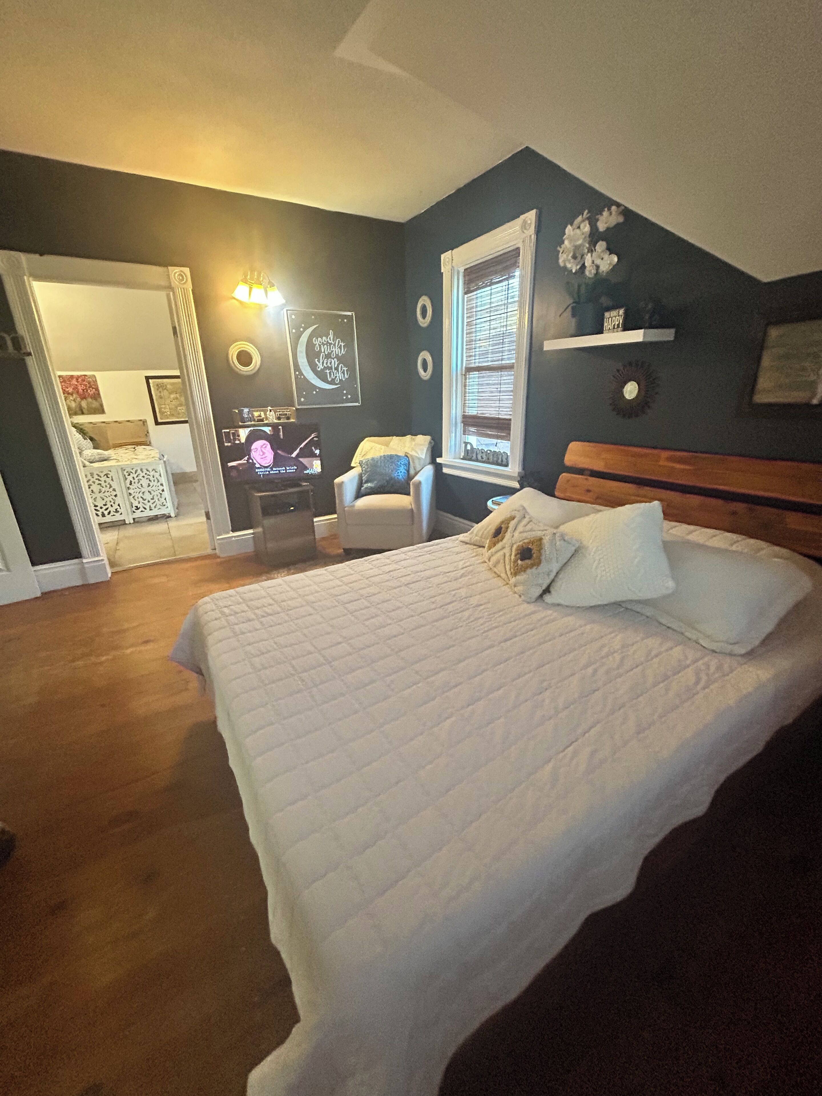 A bedroom with a large bed, a chair, a desk with a computer, and a window with blinds.