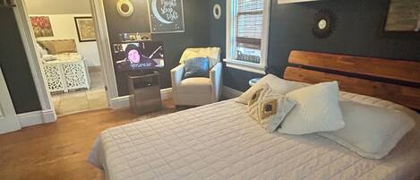 A bedroom with a large bed, a chair, a desk with a computer, and a window with blinds.