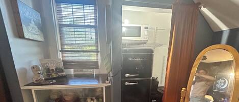 Une pièce compacte comprenant un coin cuisine, une petite fenêtre avec stores, un meuble blanc avec tiroirs et un miroir avec un cadre décoratif.