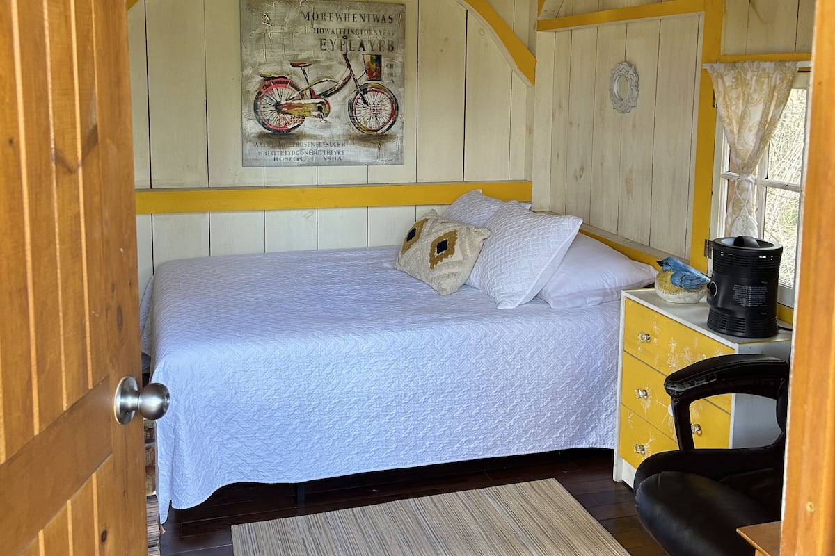 A cozy room with a bed, a desk, a chair, a window, and a wall-mounted artwork.