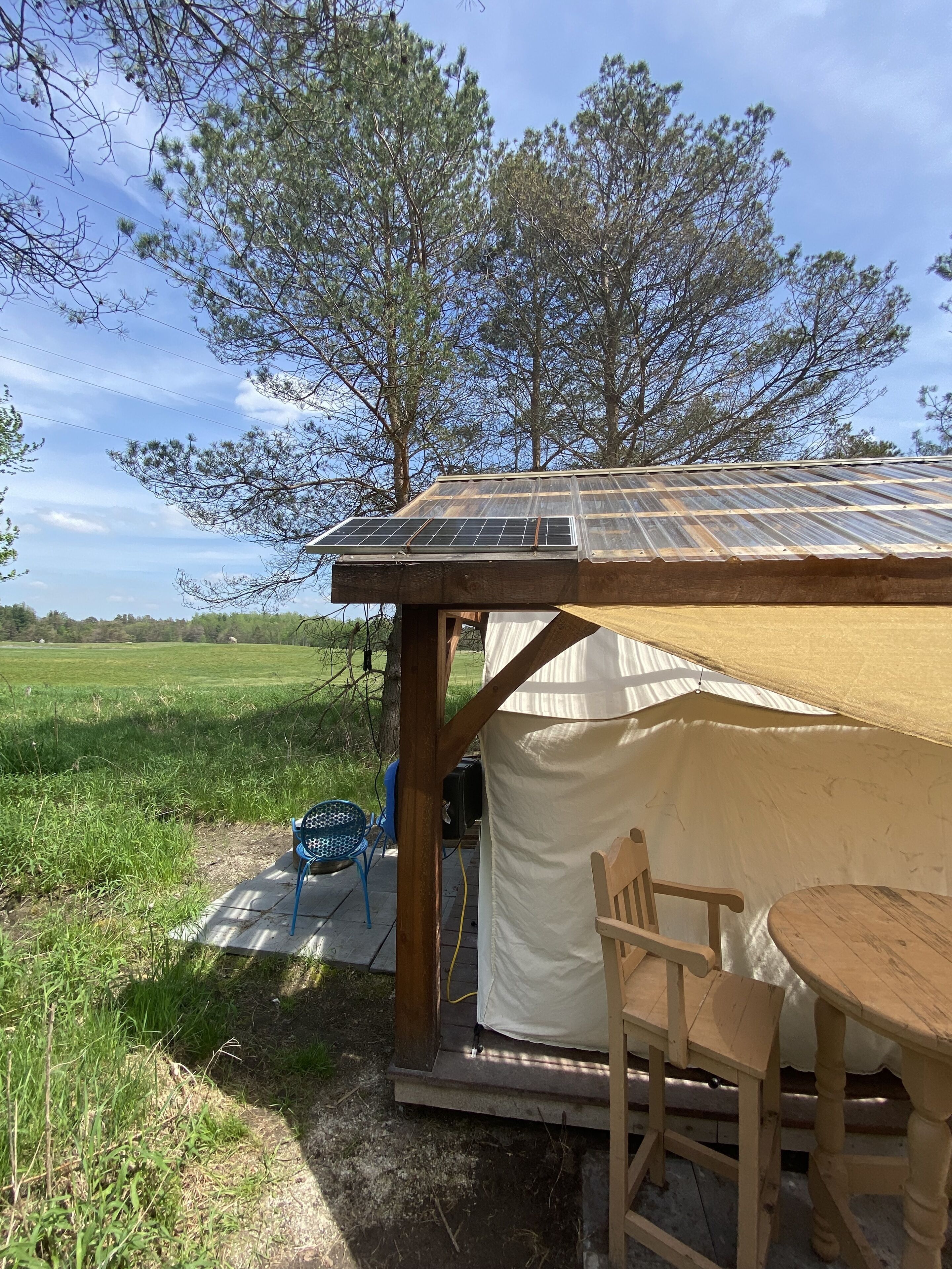 A small, open-sided structure with a solar panel roof, a tent-like interior, and a table and chair outside.