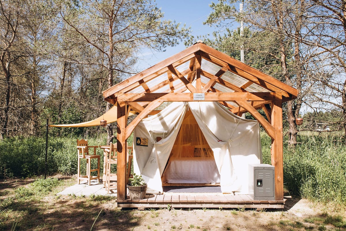 A wooden cabin with a pitched roof, a small porch with chairs, and a bed inside.