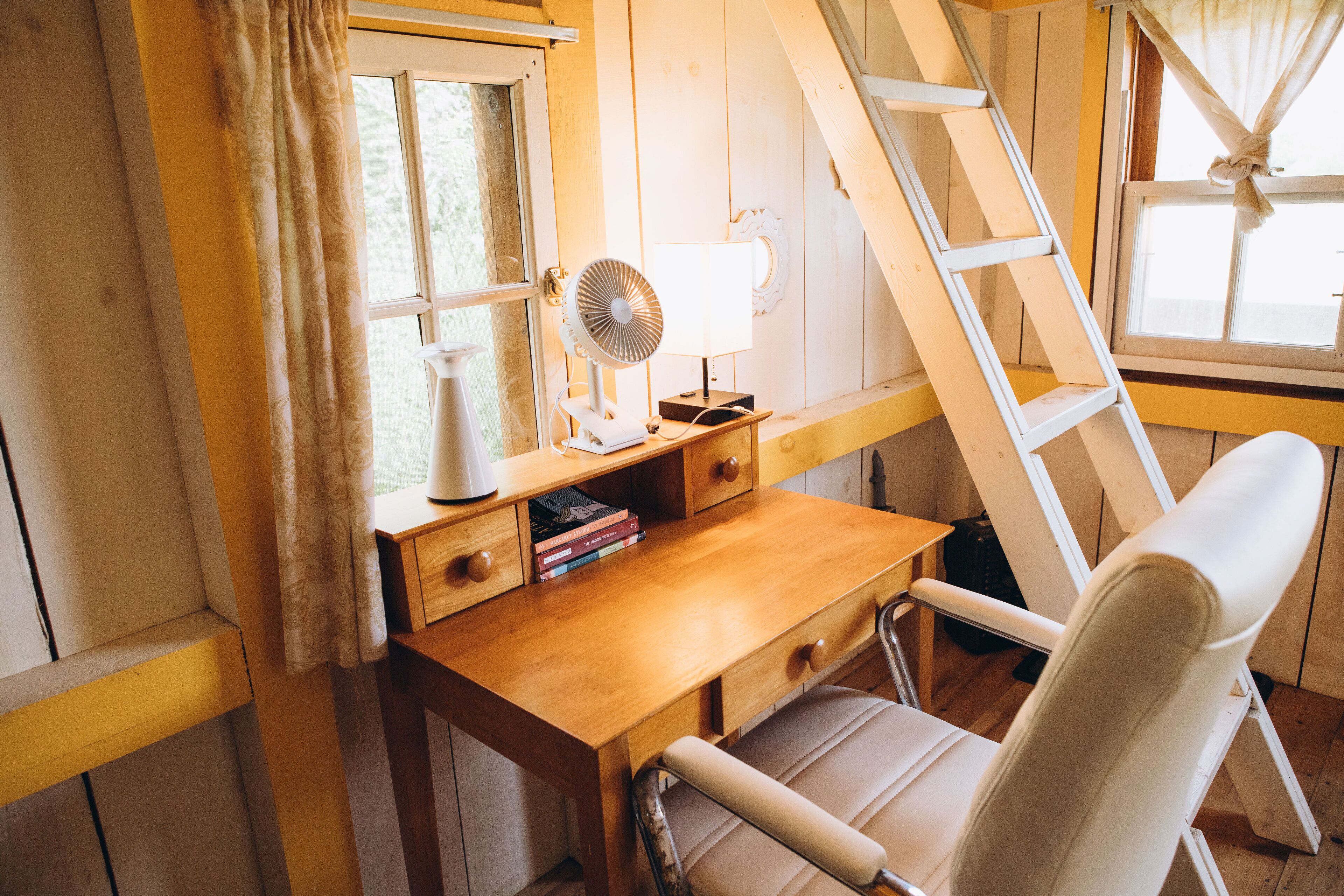A wooden desk with a white chair, a standing fan, and a window with curtains.