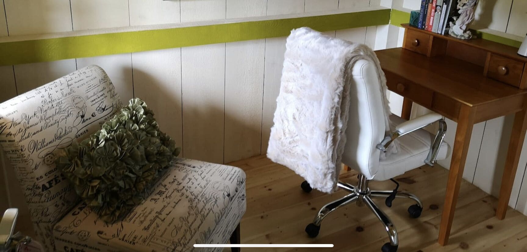 Una stanza con una sedia bianca rivestita da una coperta bianca, una scrivania in legno e una libreria piena di libri e oggetti decorativi.