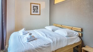 Una cama bien hecha con sábanas blancas y una toalla enrollada, un cabecero de madera, una mesita de noche pequeña y un cuadro en la pared.