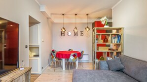 Una sala de estar moderna con un sofá gris, una mesa de comedor con sillas de colores, una estantería y un televisor sobre un mueble de madera.