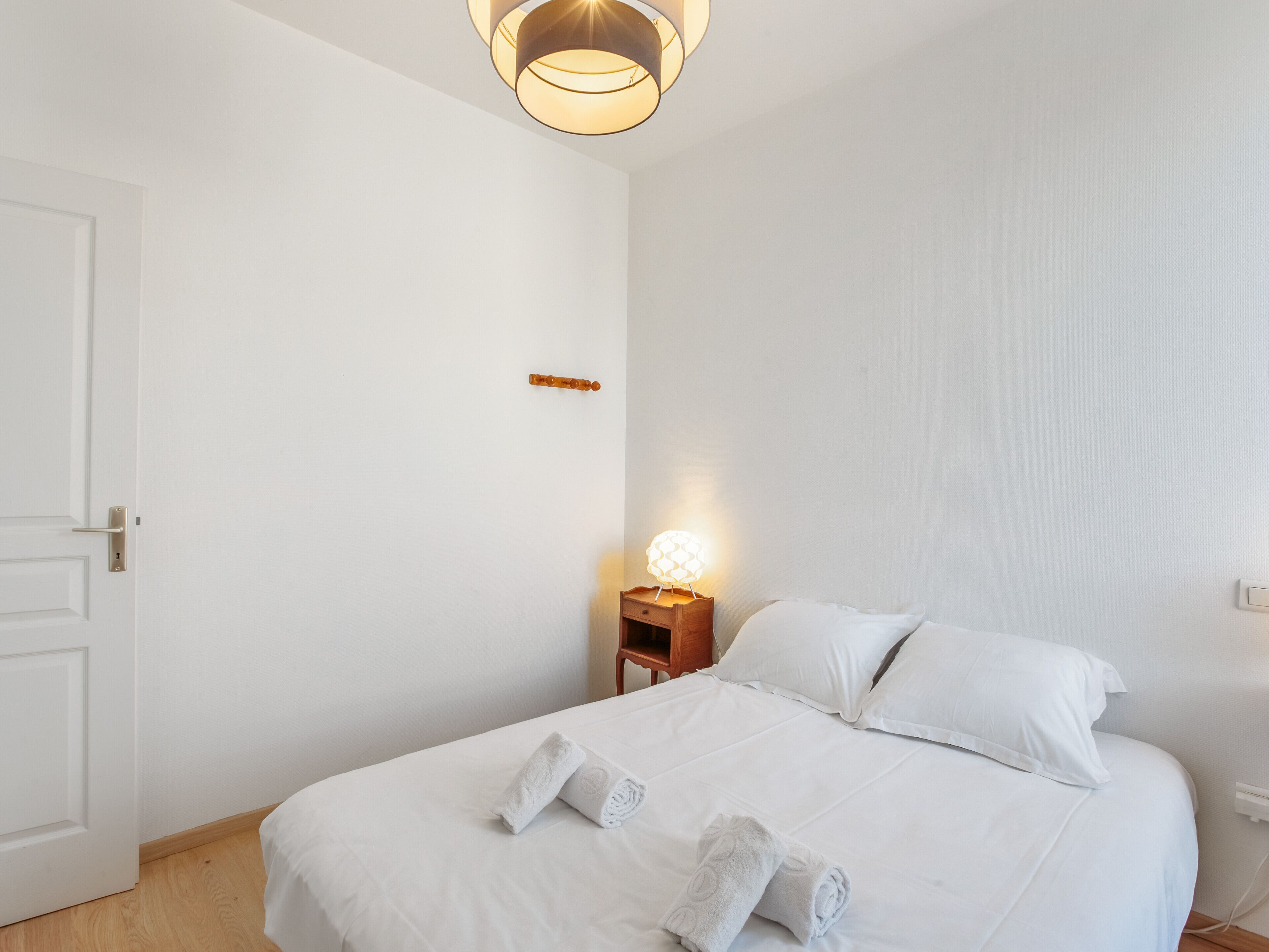Una cama bien tendida con sábanas blancas y toallas enrolladas, una lámpara de noche y una mesita de madera.