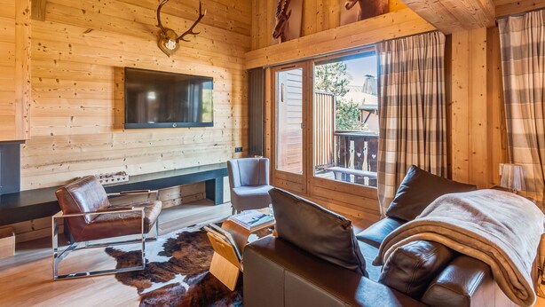 A wooden cabin interior with a TV mounted on the wall, leather sofas, a fireplace, and wooden ceiling beams. - Appartement Bridan - Welkeys (Megève)