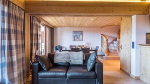 A cozy living room with a leather sofa, a dining area with a table and chairs, wooden beams on the ceiling, and a staircase leading upstairs. - Appartement Bridan - Welkeys (Megève)