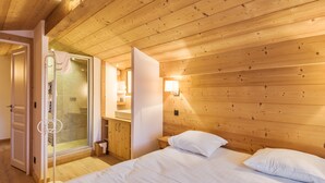 A wooden-paneled bedroom with a bed, a nightstand, a mirror, and a bathroom visible through an open door. - Appartement Bridan - Welkeys (Megève)