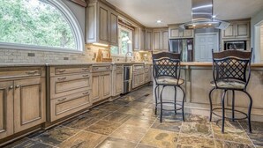 A kitchen with a large window, stone tiles, wooden cabinets, and two bar stools. - Beautiful, Large Lake House, Made for Family Fun! (Hot Springs Village)