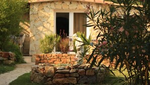 Une maison en pierre avec un toit de tuiles, un chemin de pierre menant à l’entrée et un jardin où poussent différentes espèces de plantes.