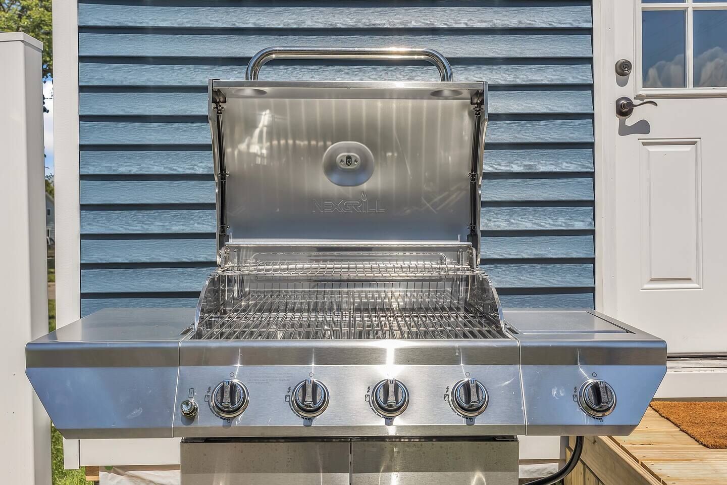 Un barbecue en acier inoxydable doté de quatre brûleurs, d’un couvercle et d’une poignée, placé devant une maison dont le revêtement extérieur est bleu.