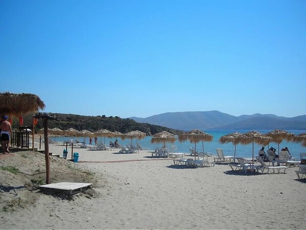 Une plage avec des chaises longues et des parasols, une terrasse en bois et une vue dégagée sur la mer et les montagnes en arrière-plan.