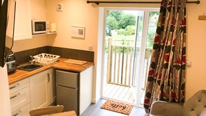 Une cuisine compacte équipée d’un four à micro-ondes, d’un évier et d’un petit réfrigérateur. On peut apercevoir une table en bois et une chaise rembourrée. Une porte-fenêtre coulissante donne accès à un espace extérieur doté d’une terrasse en bois.