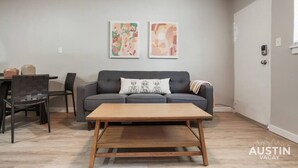 Un salón con un sofá gris, una mesa de centro de madera y dos cuadros abstractos en la pared.