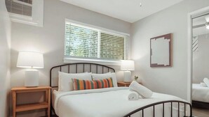Un dormitorio con cama, dos mesitas de noche, una ventana con persianas, un ventilador de techo y un espejo en la pared.