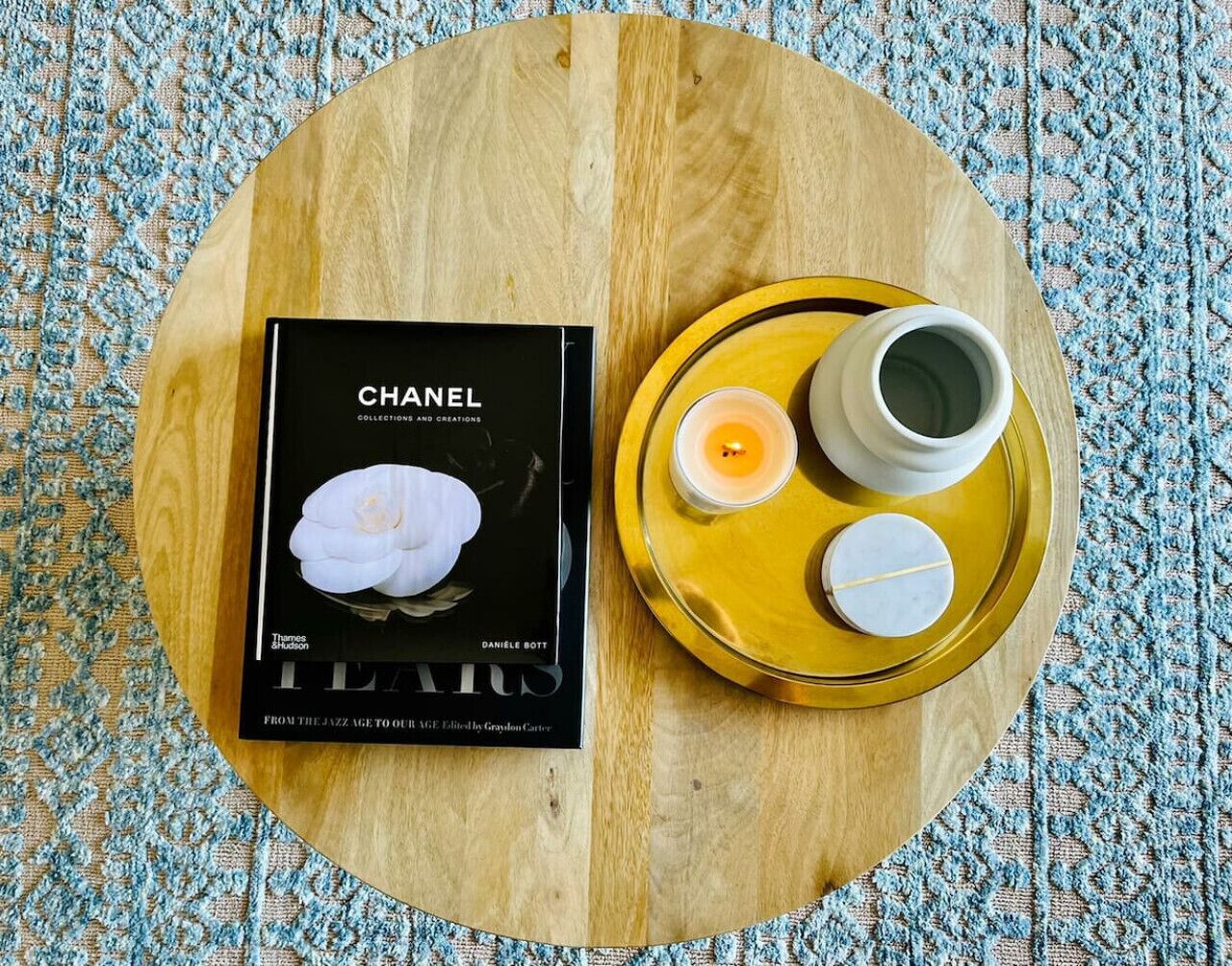 A book titled "CHANEL" on a round wooden table with a tray holding two cups and a candle.