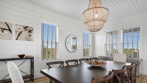 A dining area with a wooden table, six chairs, a chandelier, and large windows with blinds. - The Belvedere - Luxury Vacation Rental Near the Beach with Huge Pool & Volleyball Court (Santa Rosa Beach)