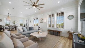 A spacious living room with a high ceiling fan, a dining area with a wooden table and chairs, and large windows with shutters. - The Belvedere - Luxury Vacation Rental Near the Beach with Huge Pool & Volleyball Court (Santa Rosa Beach)