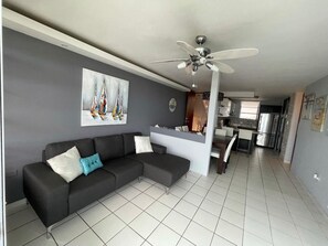 A modern living room with a sectional sofa, a ceiling fan, a painting of sailboats, and a kitchen area in the background.
