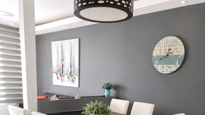 Uma área de refeições com uma mesa de madeira escura, cadeiras estofadas brancas, um vaso com planta e um relógio de parede.