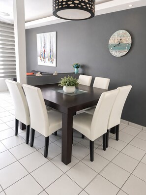 Un comedor con una mesa de madera oscura, sillas blancas tapizadas, una planta en maceta y un reloj de pared.