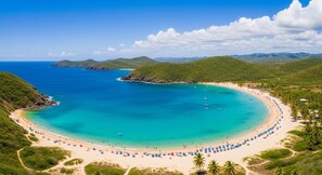 A coastal beach with clear turquoise waters, a sandy shoreline, and lush green hills in the background.