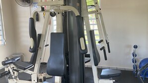 Uma academia com uma máquina de musculação multifuncional, um banco e um ventilador.