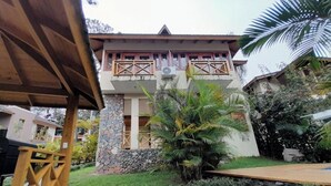 A two-story building with a stone facade, wooden balcony railings, and a small garden area with palm trees and shrubs.