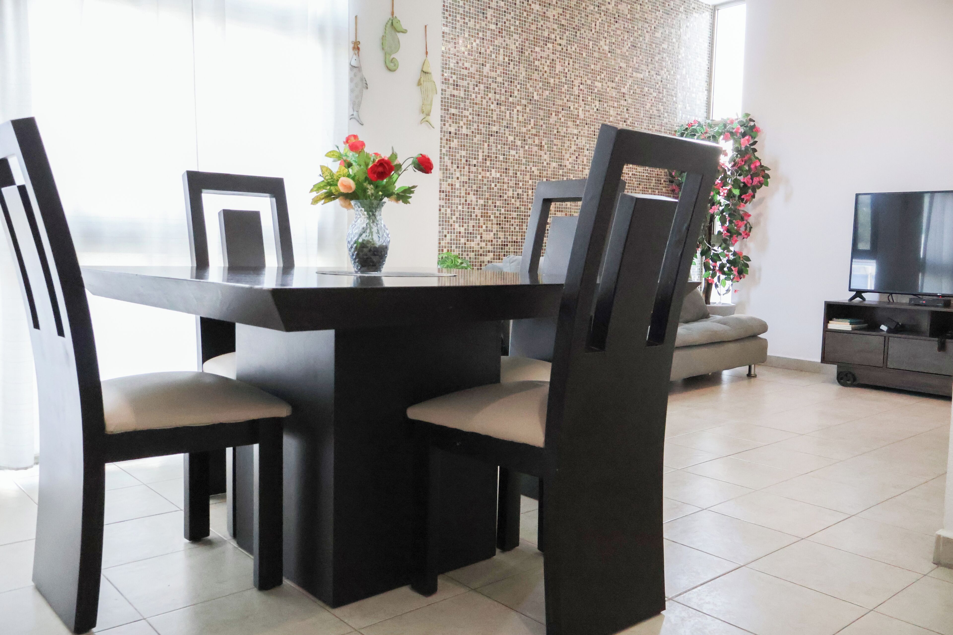 A dining area with a black table and chairs, a vase with flowers, and a television in the background.