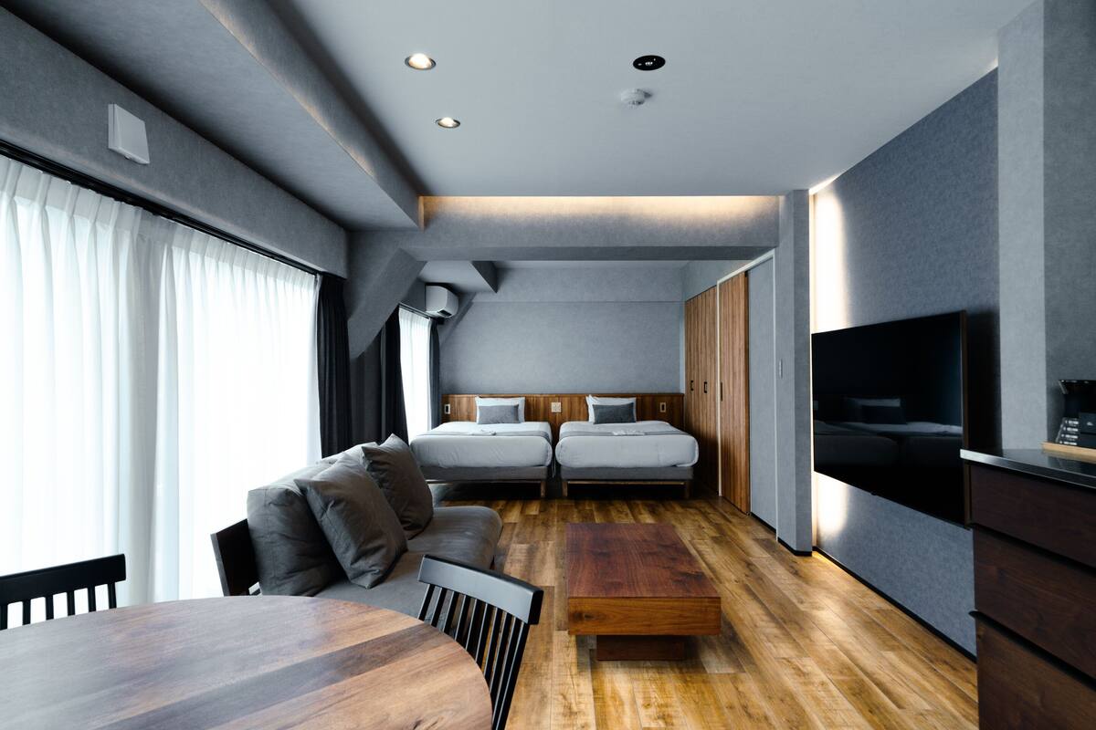 A modern hotel room with a wooden dining table, two beds, a sofa, and a TV.